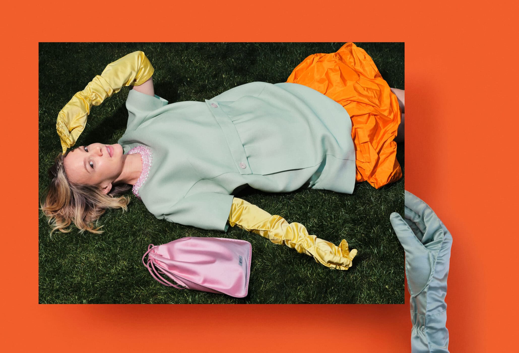 Woman in vintage fashion dress lies on a green lawn, wearing yellow gloves, near an orange cloth and pink purse in an editorial shot.