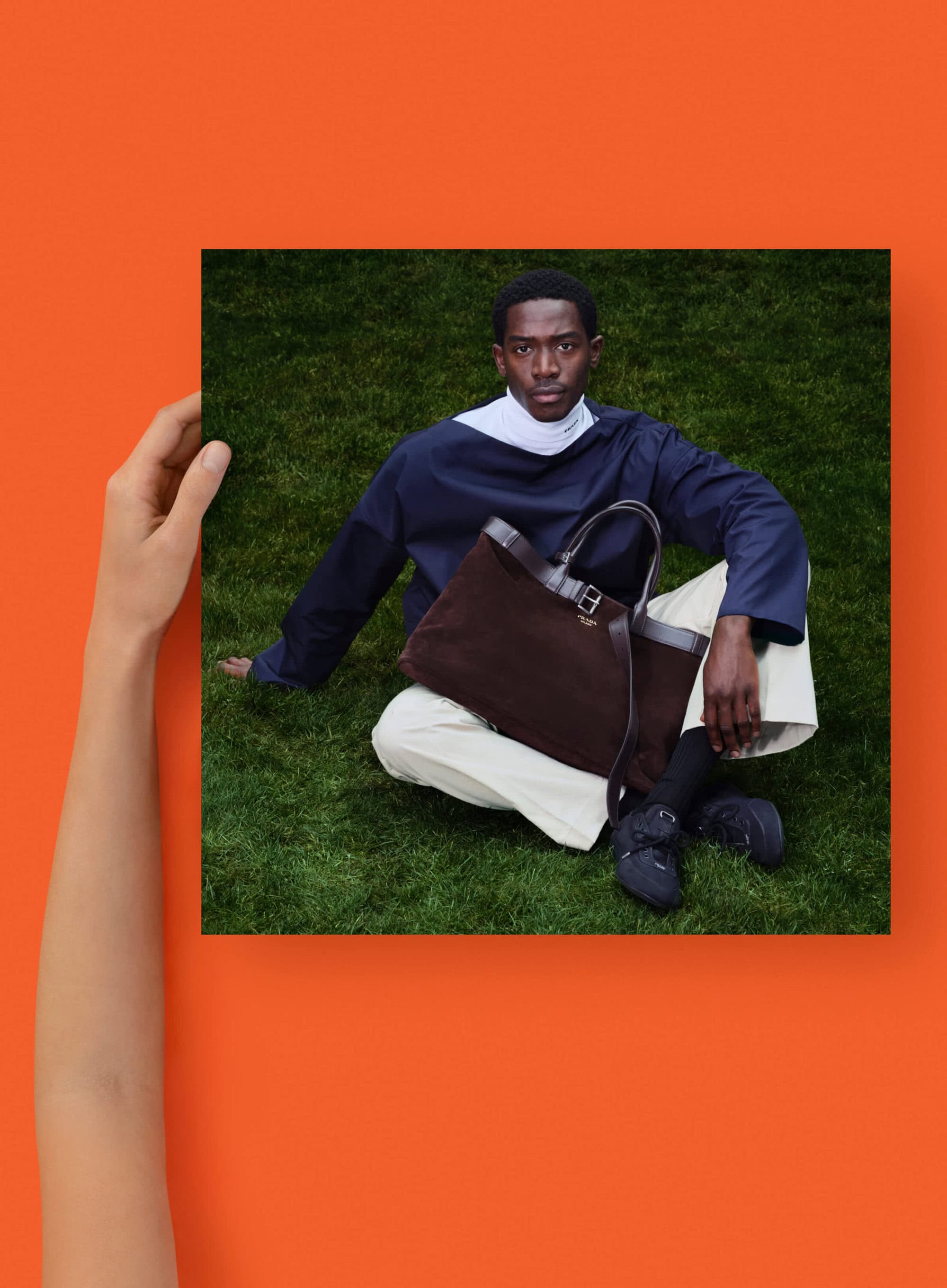Portrait of an African American man in a navy shirt and beige pants sitting on grass, next to a brown suede tote bag.