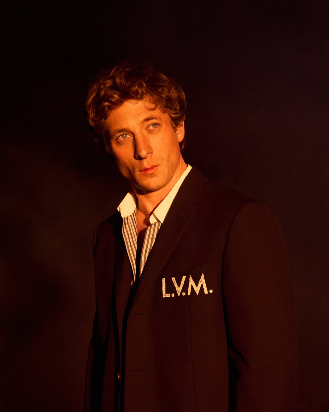 Jeremy Allen White wearing a dark suit with L.V.M. lettering and a striped shirt against a dark background.