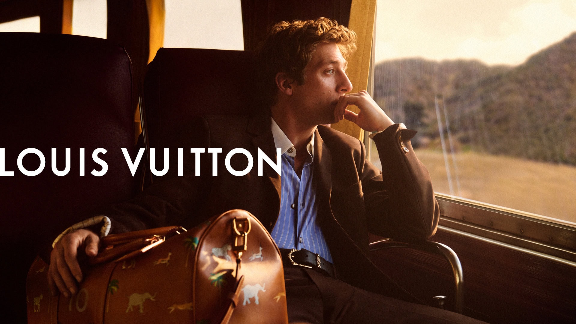 Man in suit travels by train with Louis Vuitton bag, gazing out window at landscape, evoking high fashion and luxury travel.