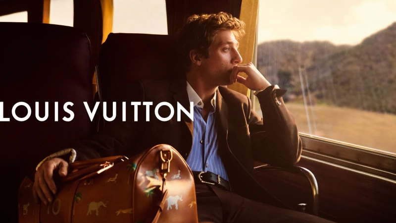 Man in suit travels by train with Louis Vuitton bag, gazing out window at landscape, evoking high fashion and luxury travel.