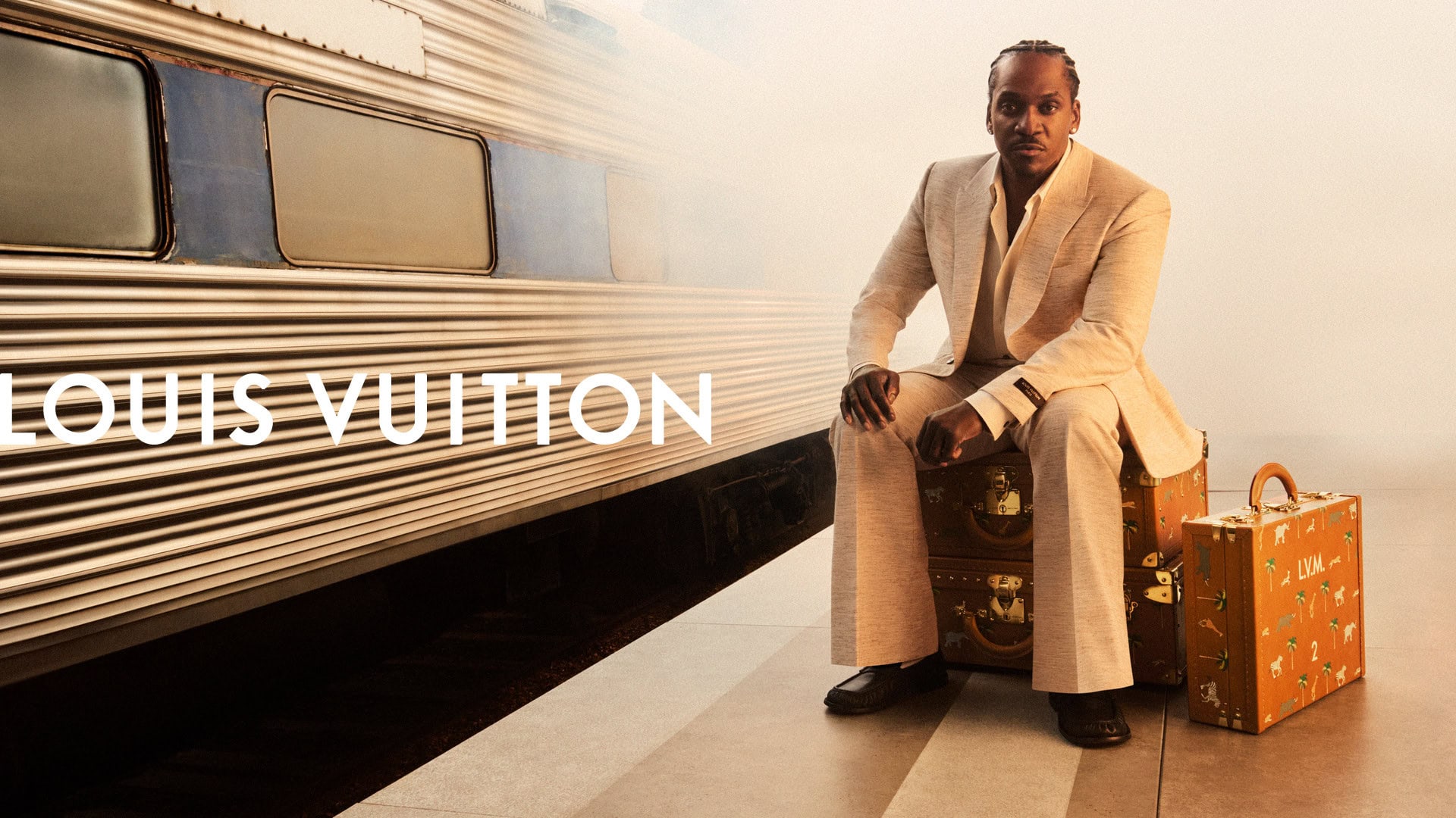 An adult male sits on stacked luggage with train in the background, wearing a suit, with the Louis Vuitton logo visible on the side.