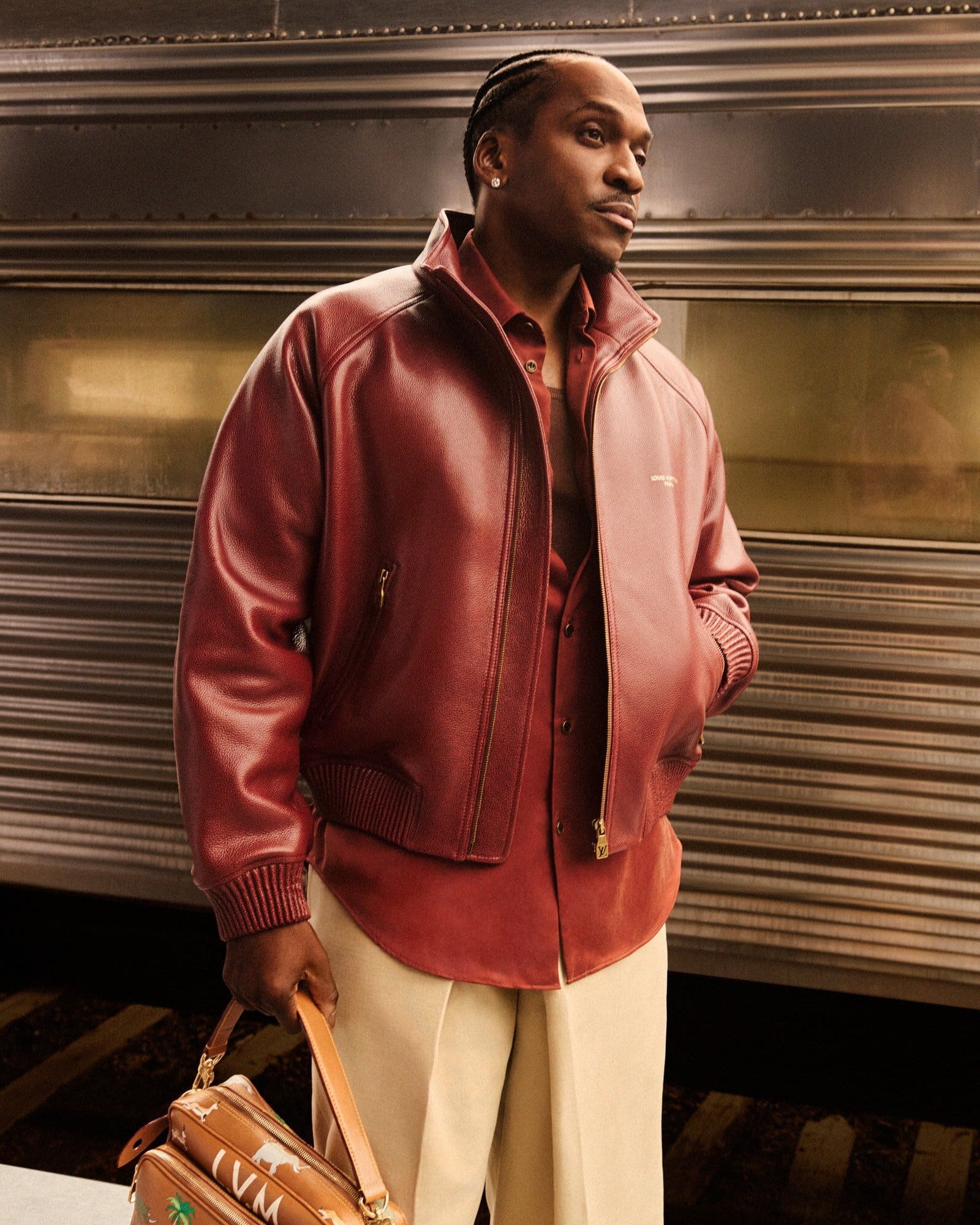 Adult male in a red leather jacket and beige trousers stands against a train with a vintage travel bag, styled for a fashion editorial.