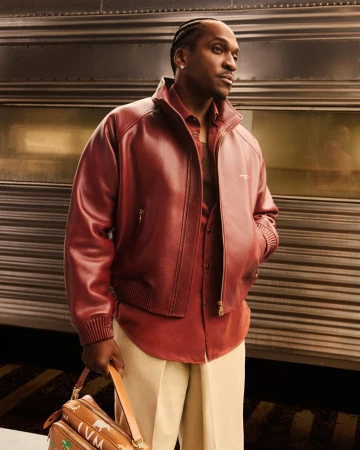 Adult male in a red leather jacket and beige trousers stands against a train with a vintage travel bag, styled for a fashion editorial.