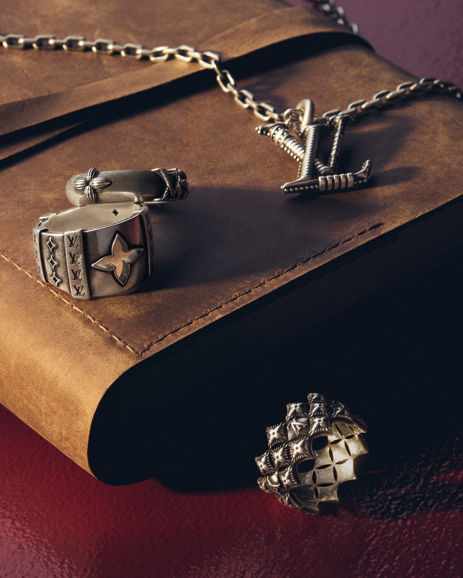 Close-up product shot of a silver-colored Louis Vuitton chain necklace, ring, and brown leather-bound notebook on a red surface.