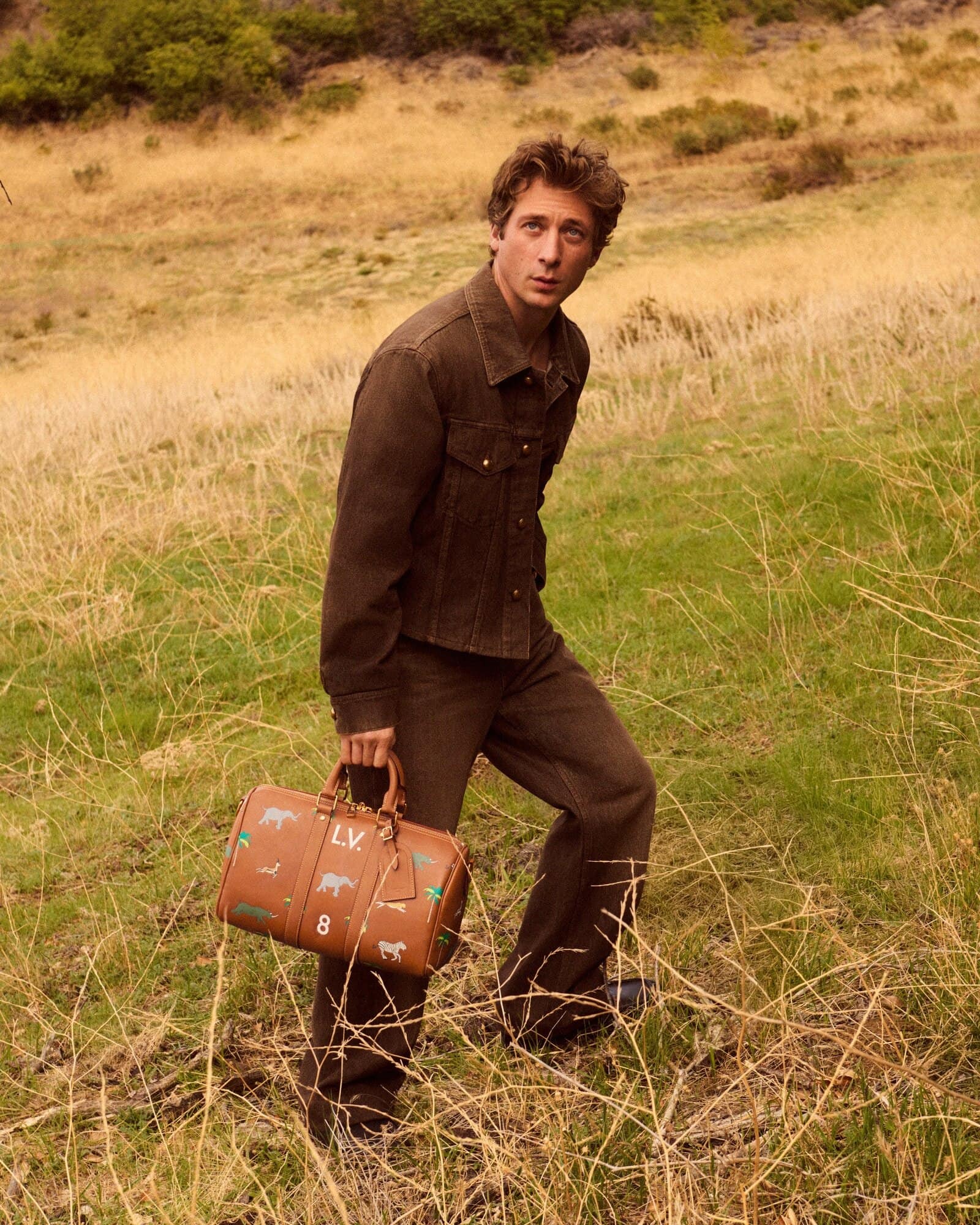 A fair-skinned man in brown jacket and trousers carrying a brown patterned satchel stands in a grassy field with brown dry grass.