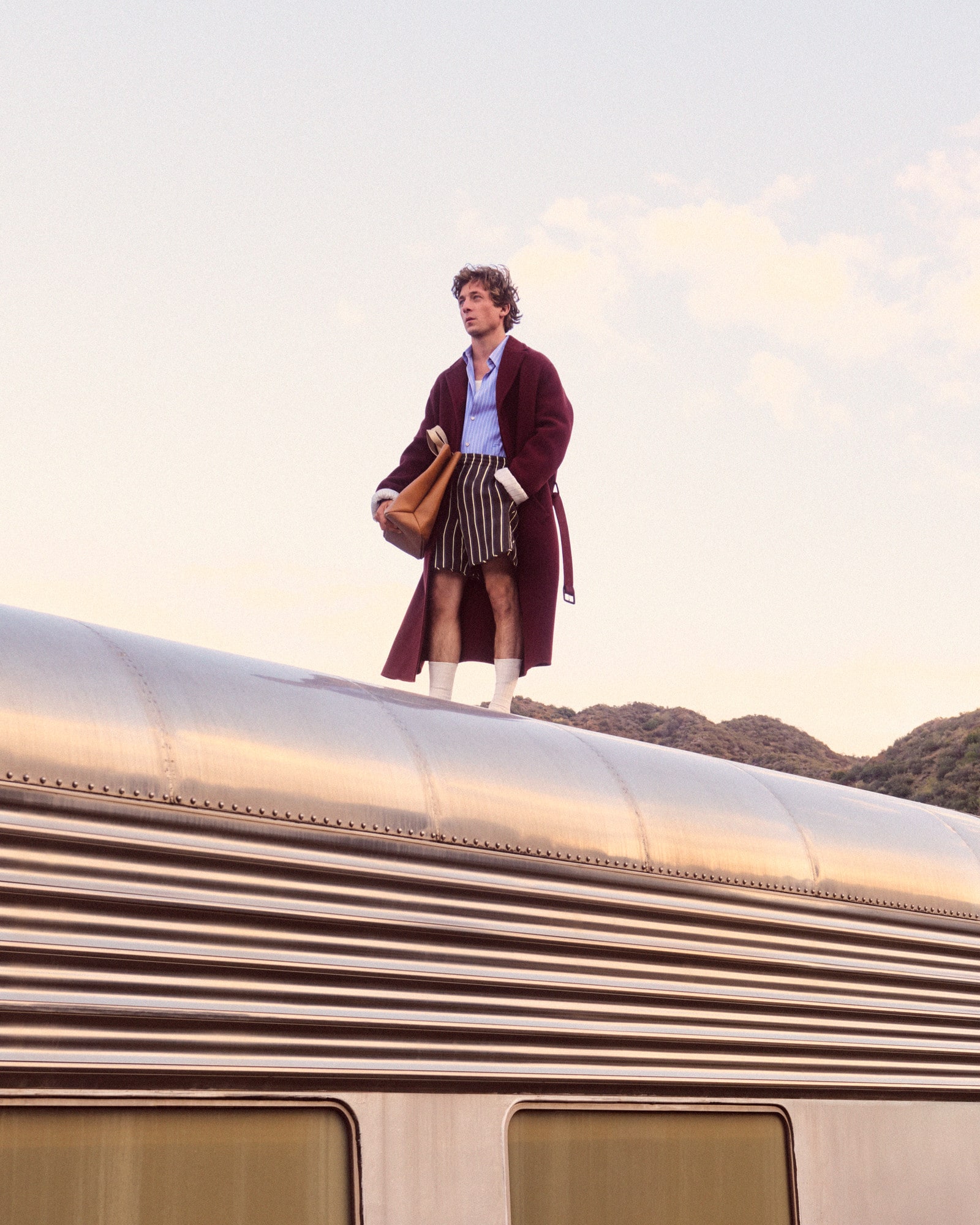 A man in a robe and striped shorts stands on the roof of a train with a tote bag, posing against a soft, pastel sky.
