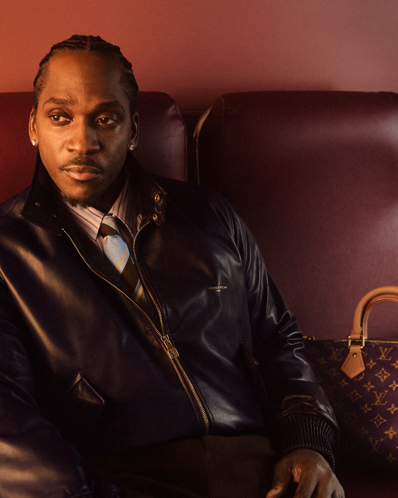 Portrait of adult Black man in blue jacket, tie, and dress shirt, seated next to a Louis Vuitton handbag on a red leather bench.