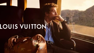 Jeremy Allen White seated inside a train next to a window, resting his hand on a brown leather duffle bag for Louis Vuitton.