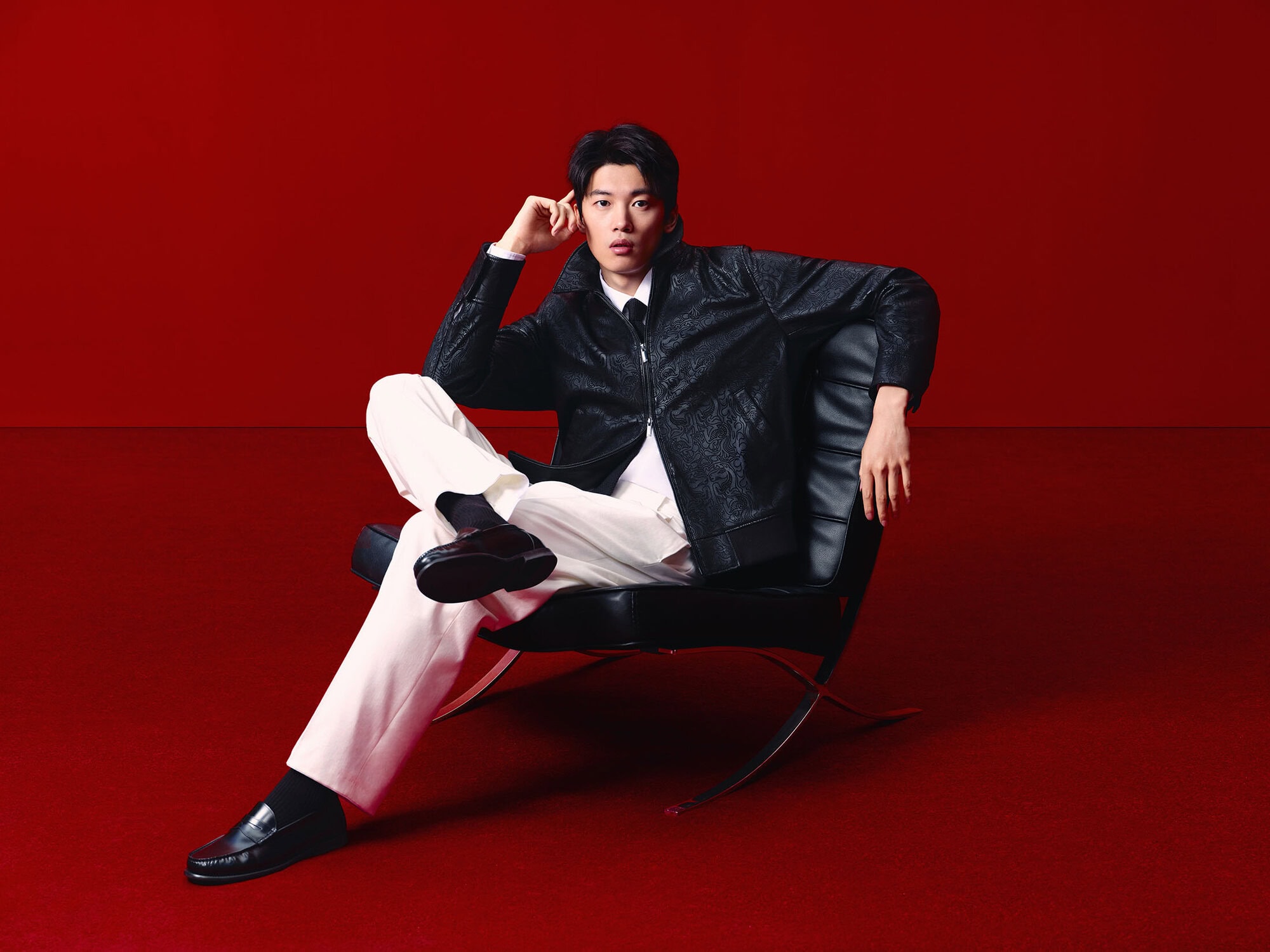 Man in a black patterned jacket and white pants poses in a black modern chair against a solid red backdrop, fashion studio shot.