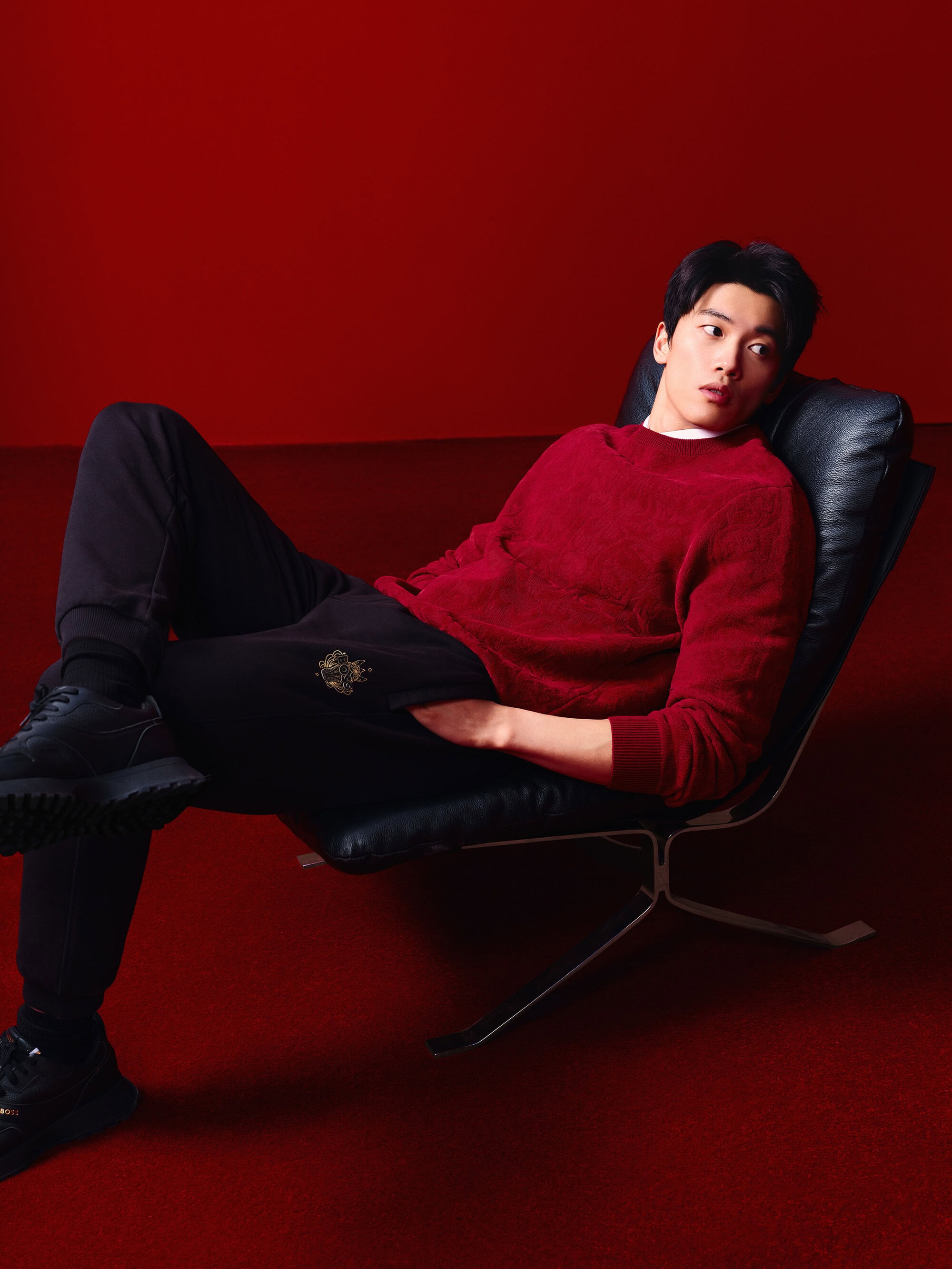 Adult male lying back on a black chair against a red backdrop. He wears a red sweater, black pants, and black shoes in studio lighting.