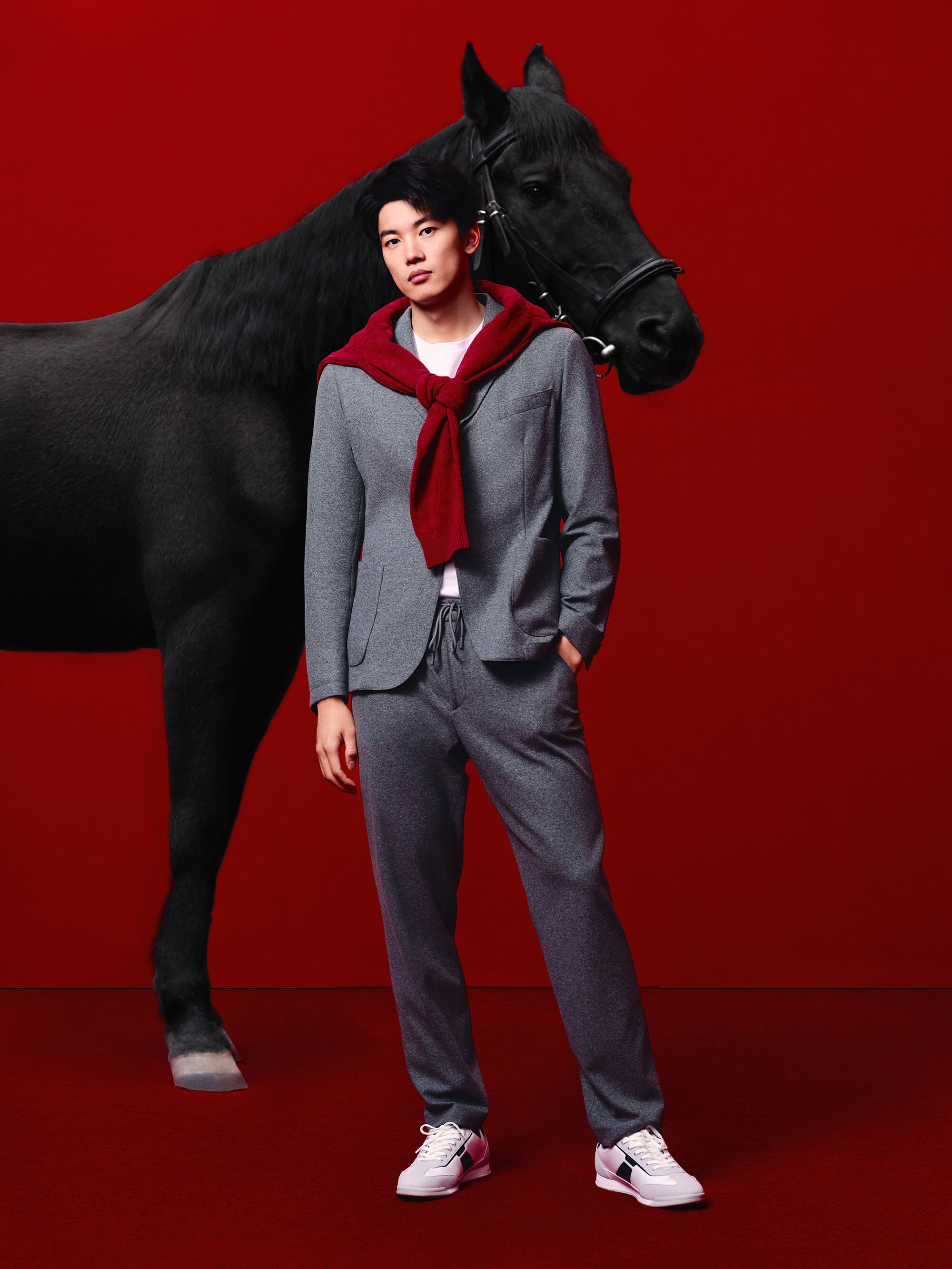 A stylish Asian man poses in a grey suit with a red sweater, alongside a sleek black horse, against a solid red background.
