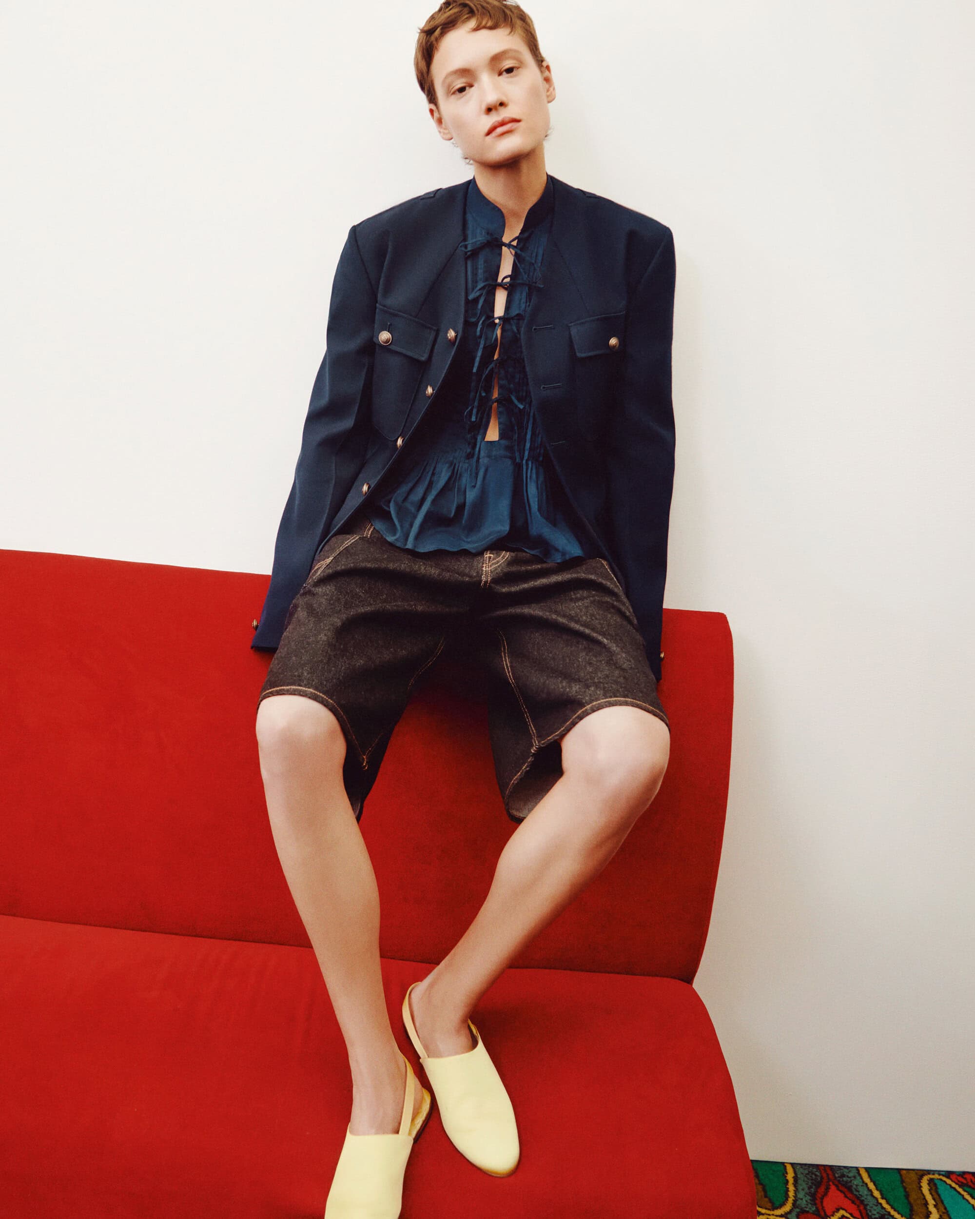 Woman seated on red couch, wearing a navy jacket and denim shorts, with bright yellow shoes, against a white wall backdrop.