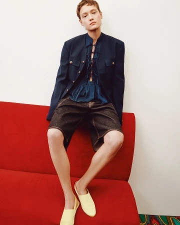 Woman seated on red couch, wearing a navy jacket and denim shorts, with bright yellow shoes, against a white wall backdrop.