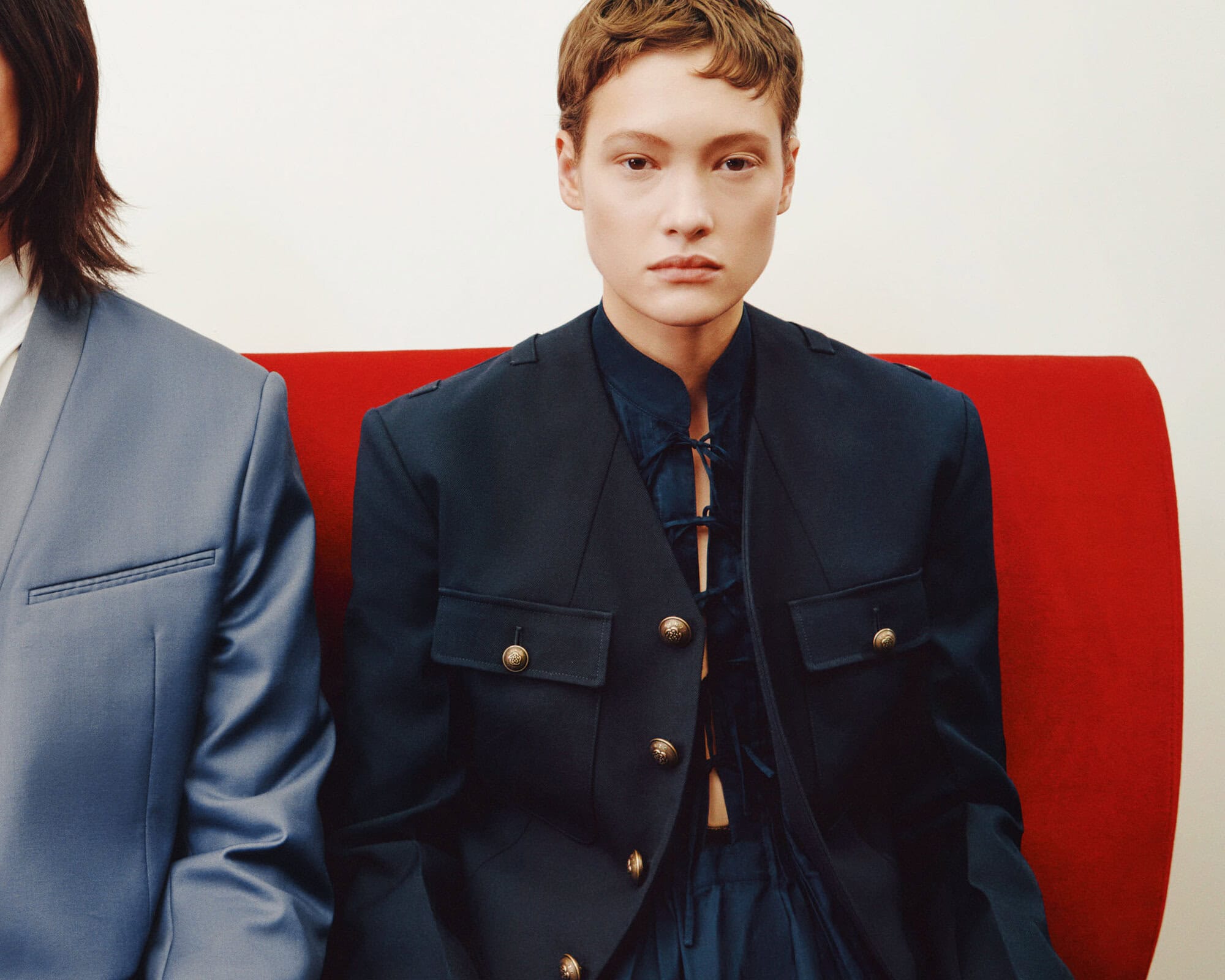 Two people sit on a red bench in navy and light blue suits. One woman gazes directly at the camera in a formal setting.