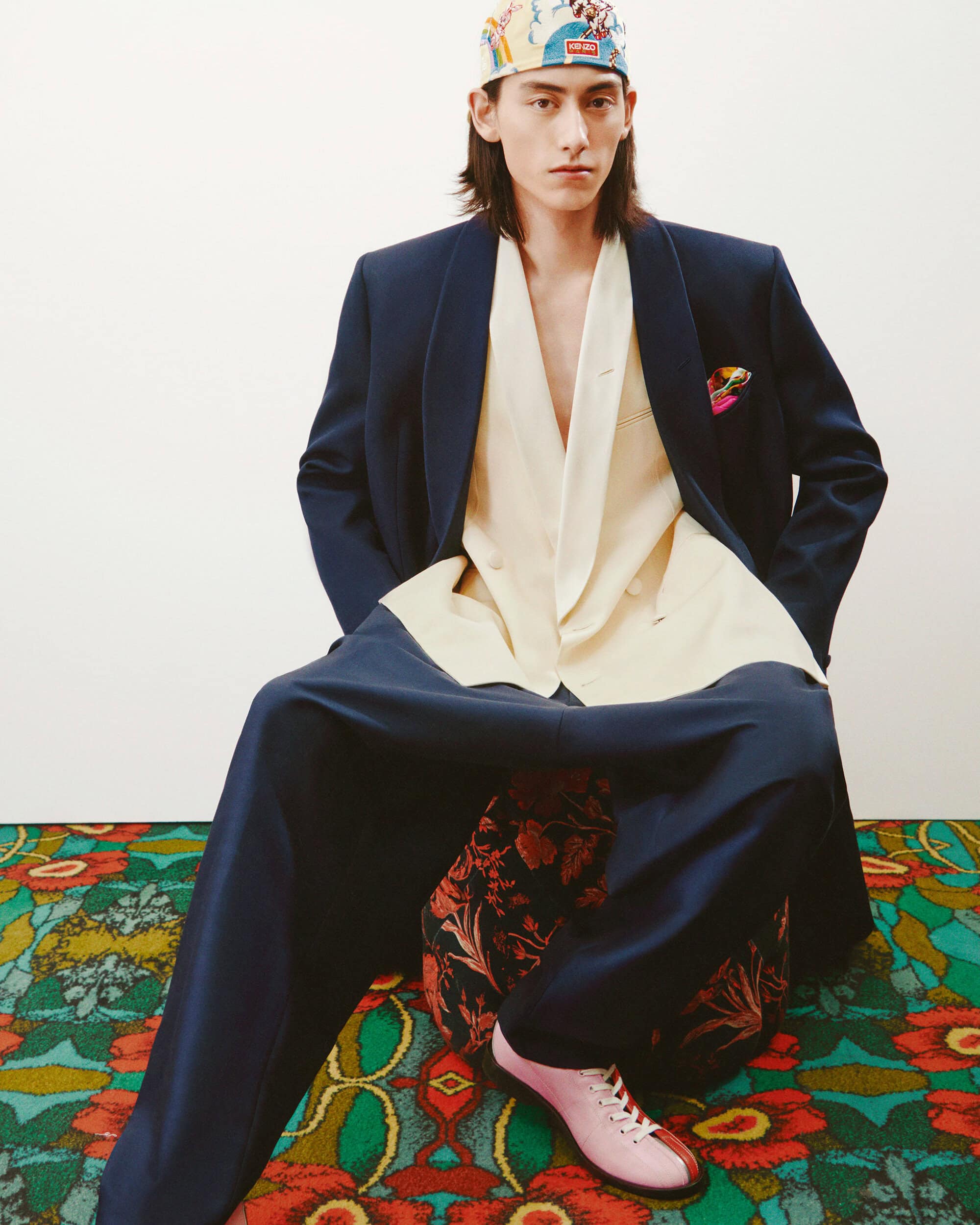 Man in blue suit and patterned headscarf poses indoors on patterned pouf. Beige blazer, pink shoes, white wall background.