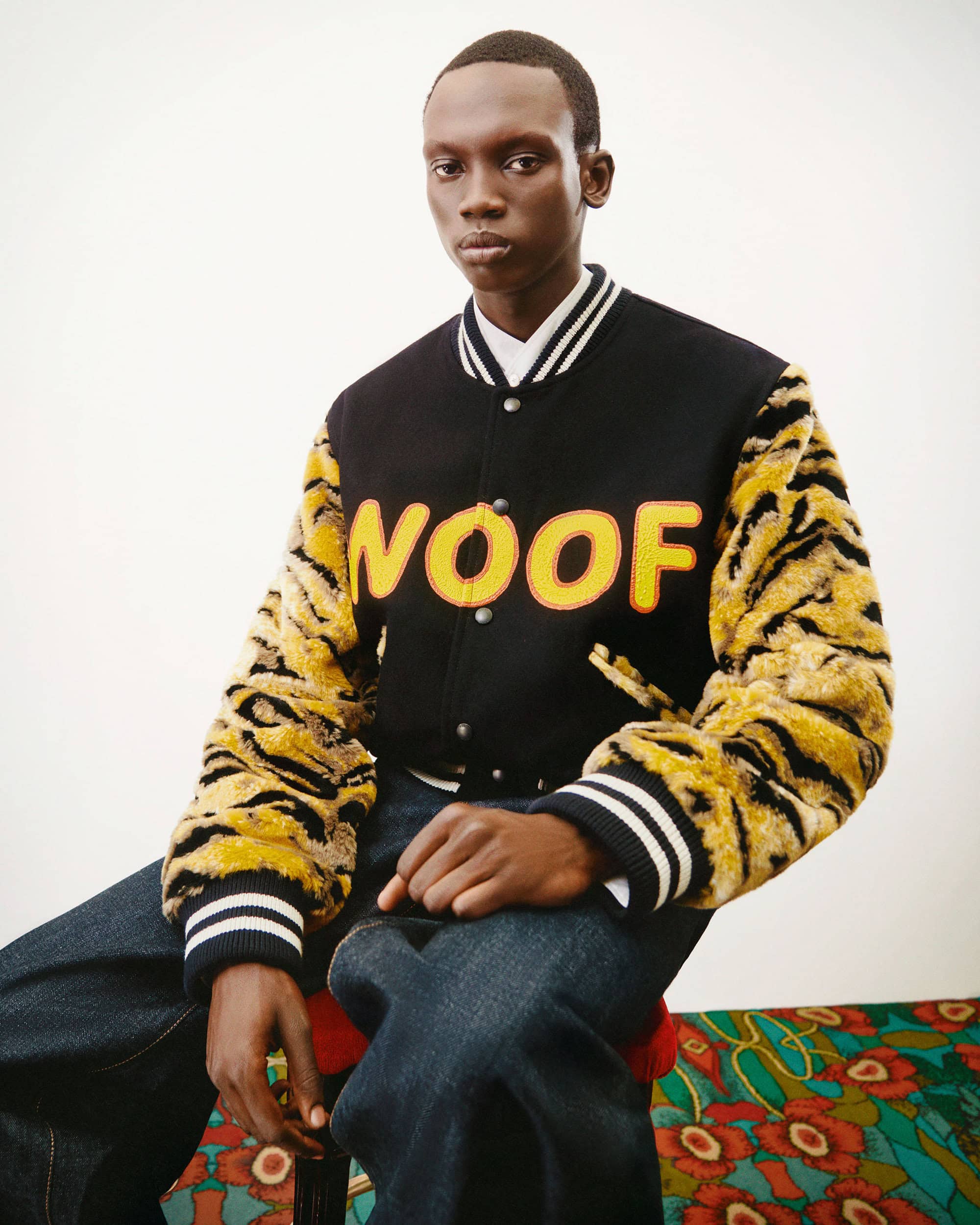 A young Black man in a varsity jacket with tiger stripe sleeves sits on a stool against a white background, looking directly at the camera.