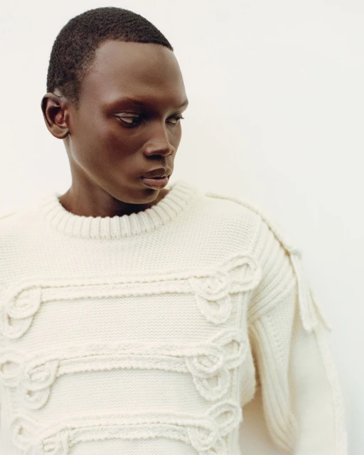 Portrait of an adult Black male wearing a textured cream-colored knit sweater, set against a neutral background in an indoor setting.
