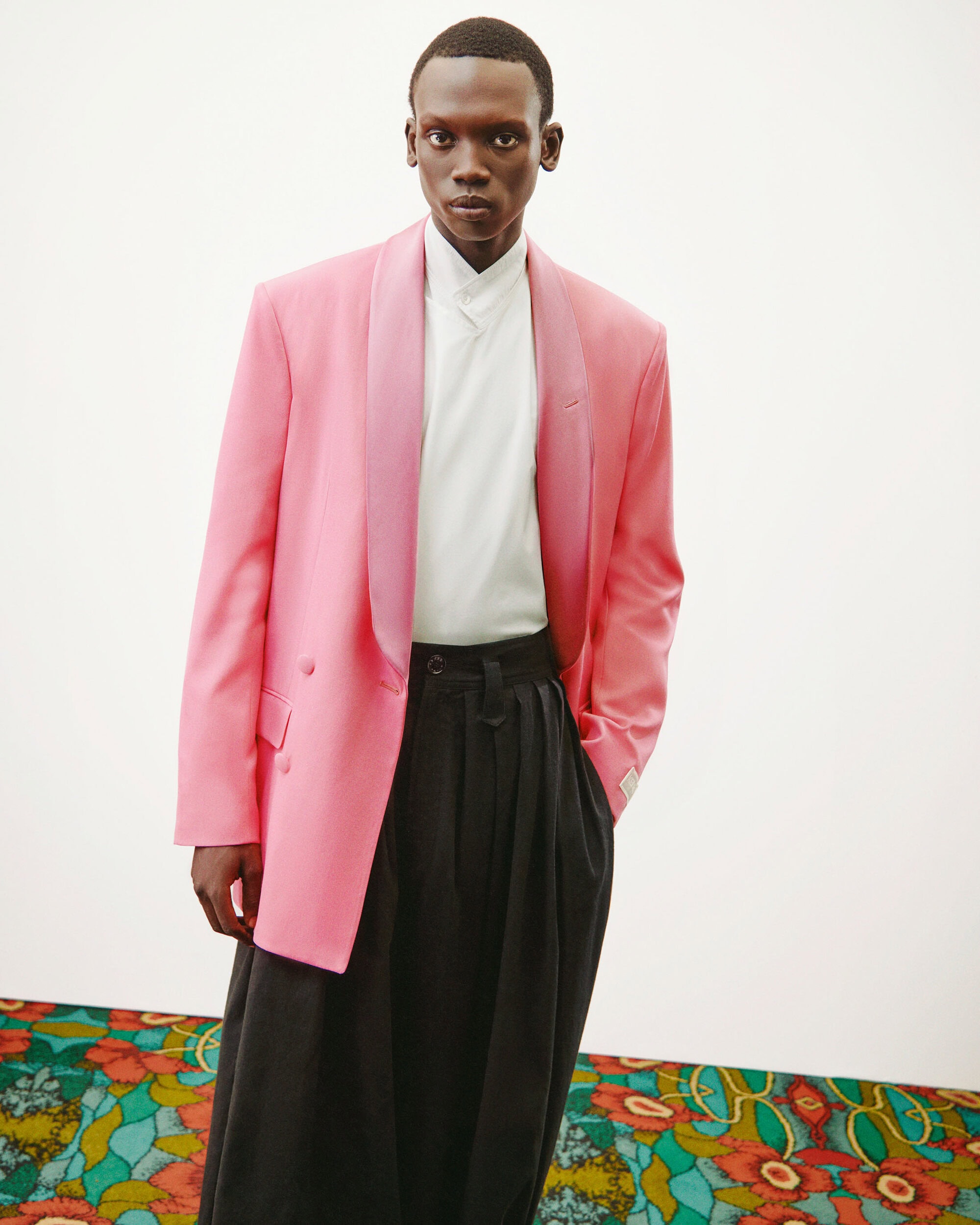 Man in a pink blazer and wide black pants stands against a white background with patterned base. Fashion editorial style portrait.