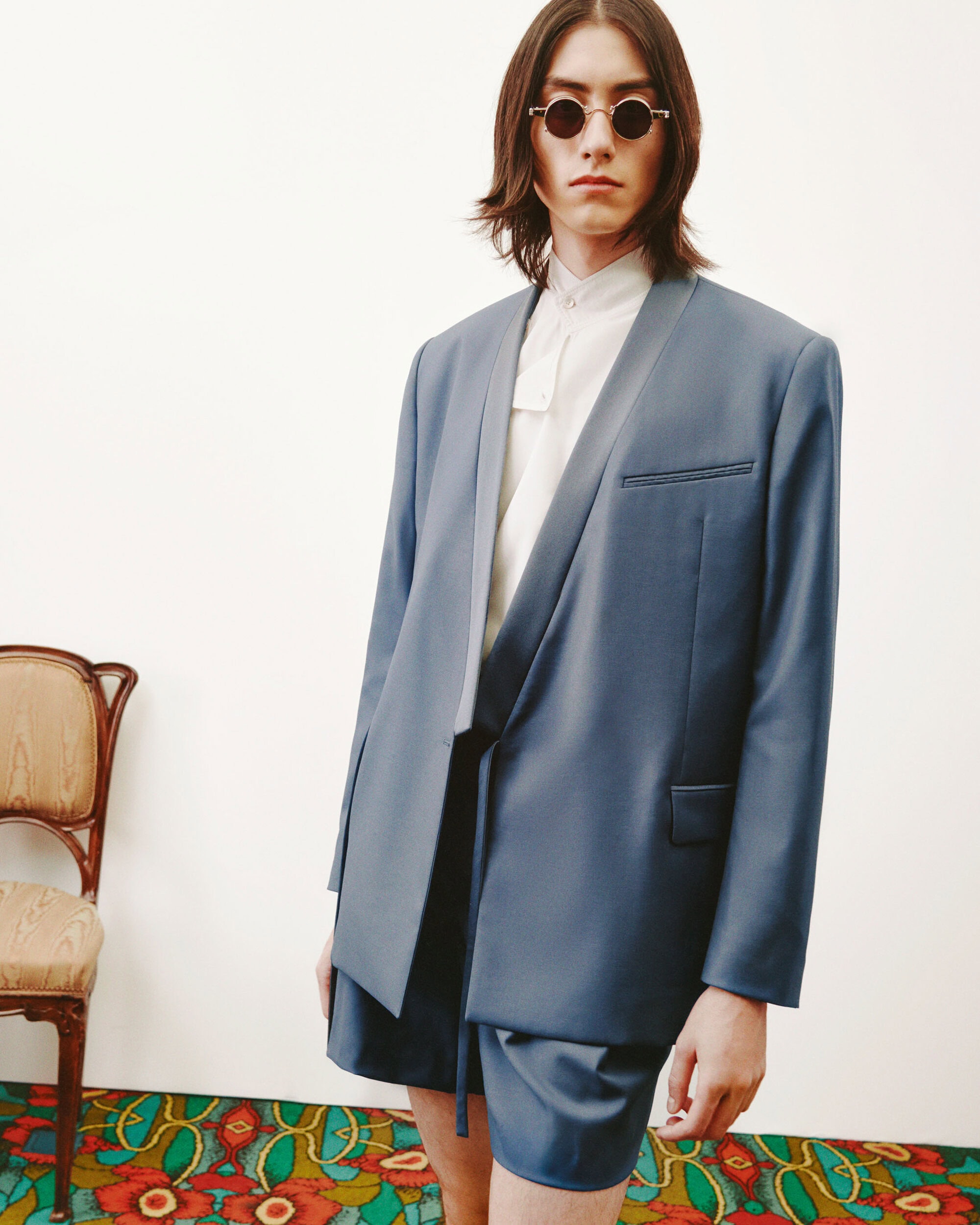 Young adult male fashion model in a blue suit jacket and shorts poses in a minimalist studio, with an ornate wooden chair and patterned floor.