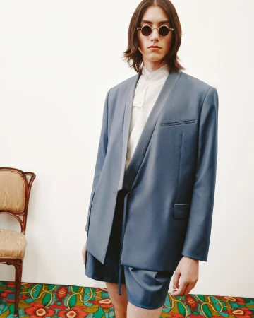 Young adult male fashion model in a blue suit jacket and shorts poses in a minimalist studio, with an ornate wooden chair and patterned floor.