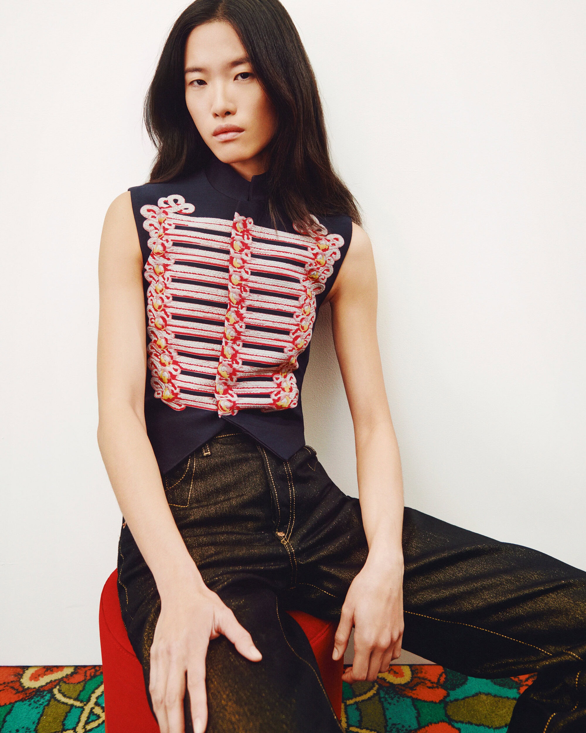 Young woman in a sleeveless top with military-style embellishments, seated on a red stool against a neutral background. Dark denim pants.