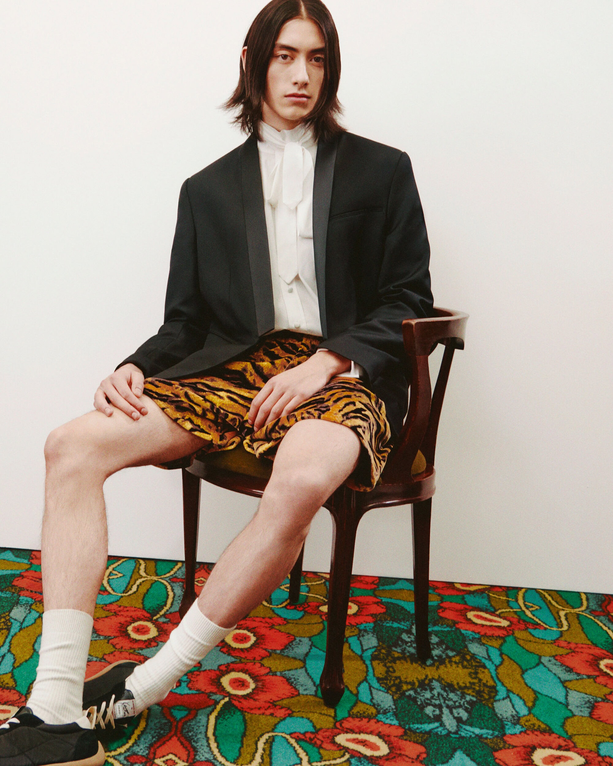 A slender man with long dark hair sits on a wooden chair wearing a blazer, blouse, and tiger-print shorts, against a white wall.