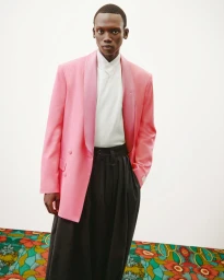 Black man wearing a pink blazer, white shirt, and dark trousers against a white studio wall