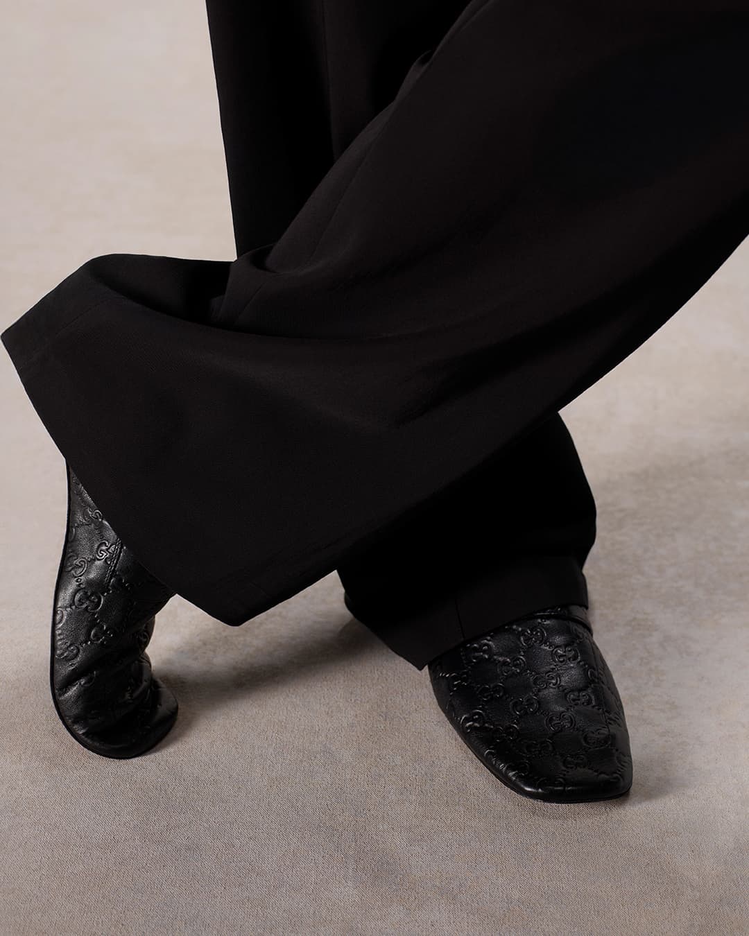 Black leather Gucci loafers with crossed legs in a studio setting show high-fashion footwear and an androgynous aesthetic.