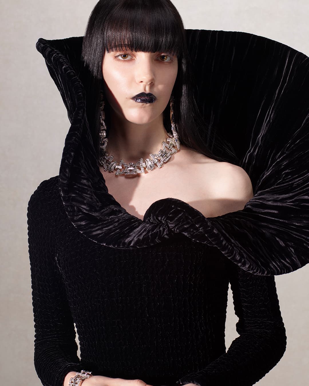 Model in black velvet high-collar dress with crystal necklace against a plain grey background