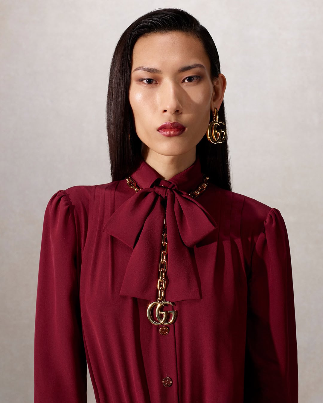 Portrait of a stylish Asian woman in a burgundy dress with a bow and gold Gucci accessories, against a light backdrop, in a fashion setting.