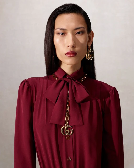 Portrait of a stylish Asian woman in a burgundy dress with a bow and gold Gucci accessories, against a light backdrop, in a fashion setting.