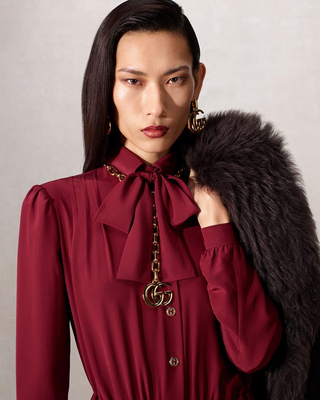 Asian model wears a burgundy dress with a bow and holds a fur coat. Gold-tone chain necklace, GG logo earrings, and pendant.