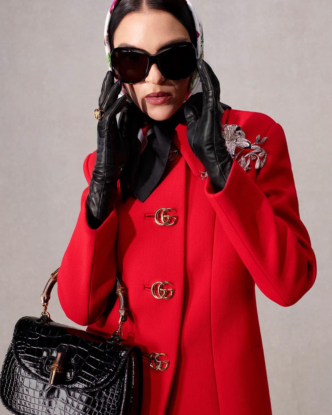 Elegant woman in a red coat, sunglasses, and headscarf carrying a black alligator purse. She's in an indoor studio.