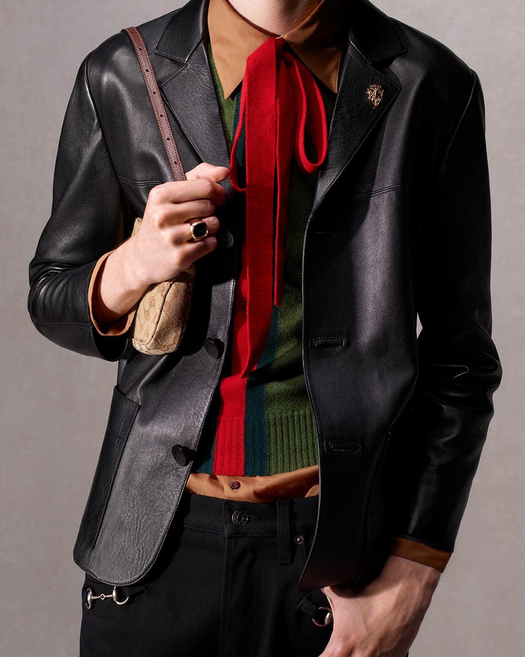 Man in a black leather jacket, tan shirt, and red tie, wearing a brown purse and black pants, standing indoors against a neutral wall.