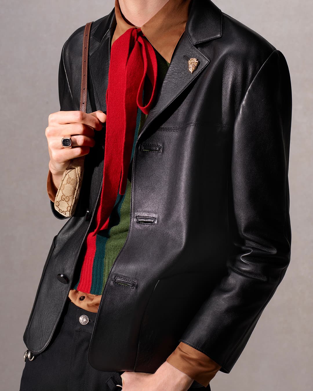 Fashionably dressed person in a black leather jacket with a colorful scarf, and a small purse with a logo print, set against a neutral backdrop.