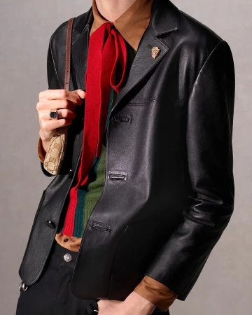 Fashionably dressed person in a black leather jacket with a colorful scarf, and a small purse with a logo print, set against a neutral backdrop.