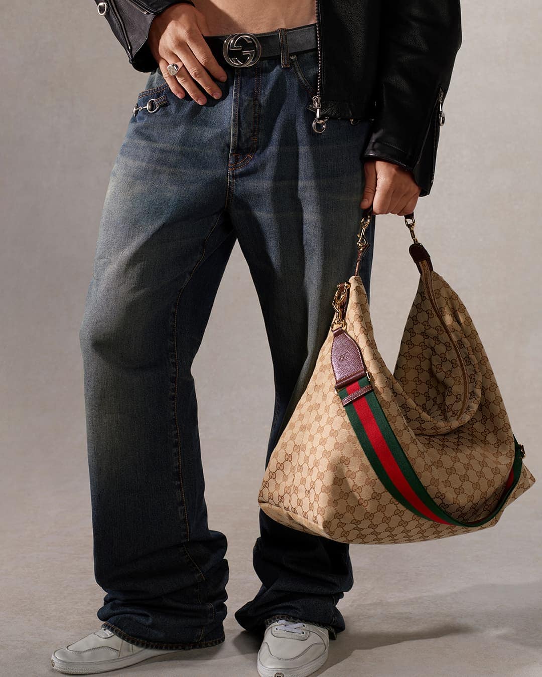 Person wearing baggy jeans, a Gucci belt, a black leather jacket, white sneakers, and holding a Gucci handbag in a studio setting.