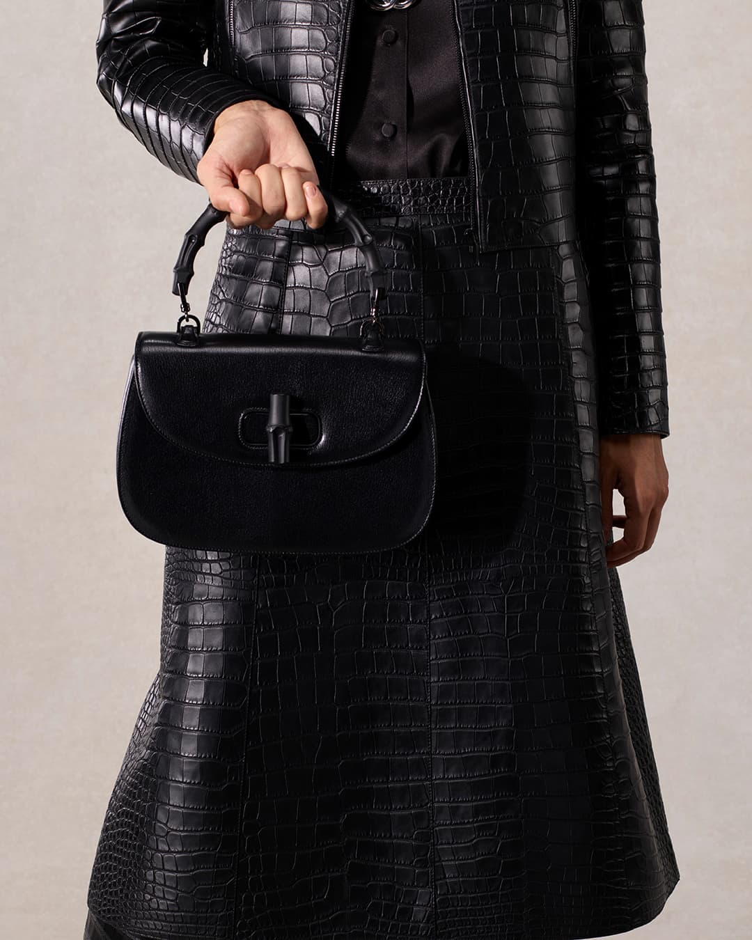 Cropped shot of a woman in a black croc-embossed outfit holding a black leather handbag with a bamboo-shaped closure.