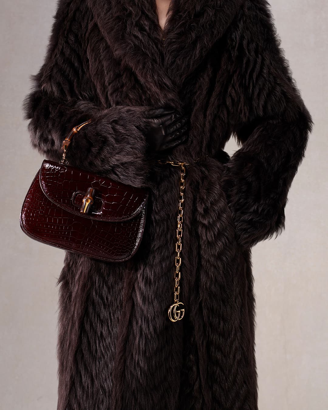 A person wearing a brown fur coat holds a burgundy crocodile handbag, accessorized with a gold chain belt with a gold logo.