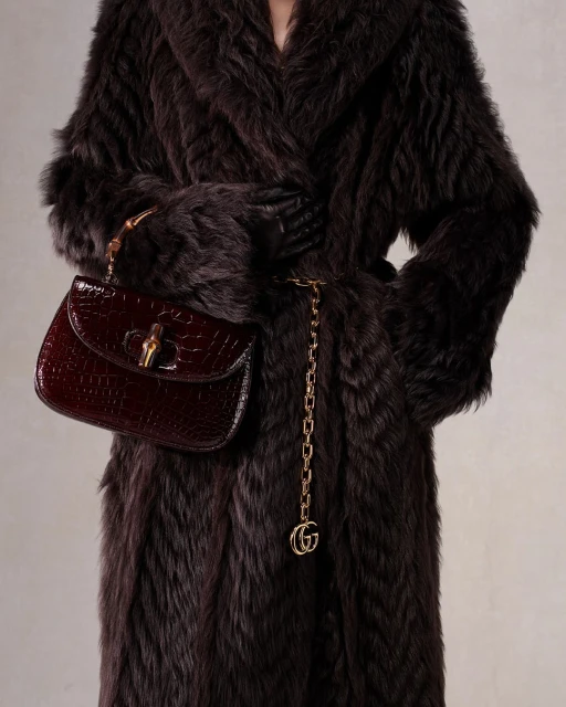 A person wearing a brown fur coat holds a burgundy crocodile handbag, accessorized with a gold chain belt with a gold logo.