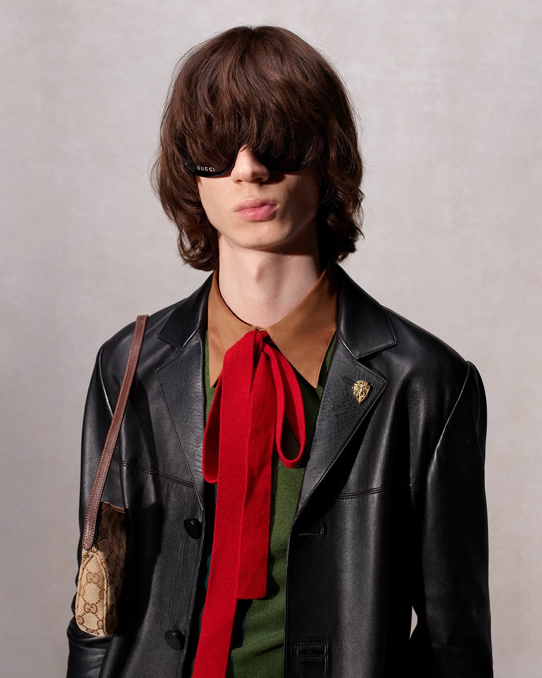 Adult male with shoulder-length hair wearing a leather jacket, glasses, and a patterned bag with a red bow tie posing indoors.