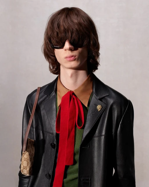 Adult male with shoulder-length hair wearing a leather jacket, glasses, and a patterned bag with a red bow tie posing indoors.
