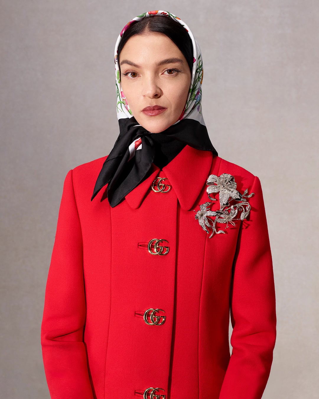 Elegant woman in a red jacket, headscarf, and ornate brooch, posing against a neutral background for a fashion editorial shot.
