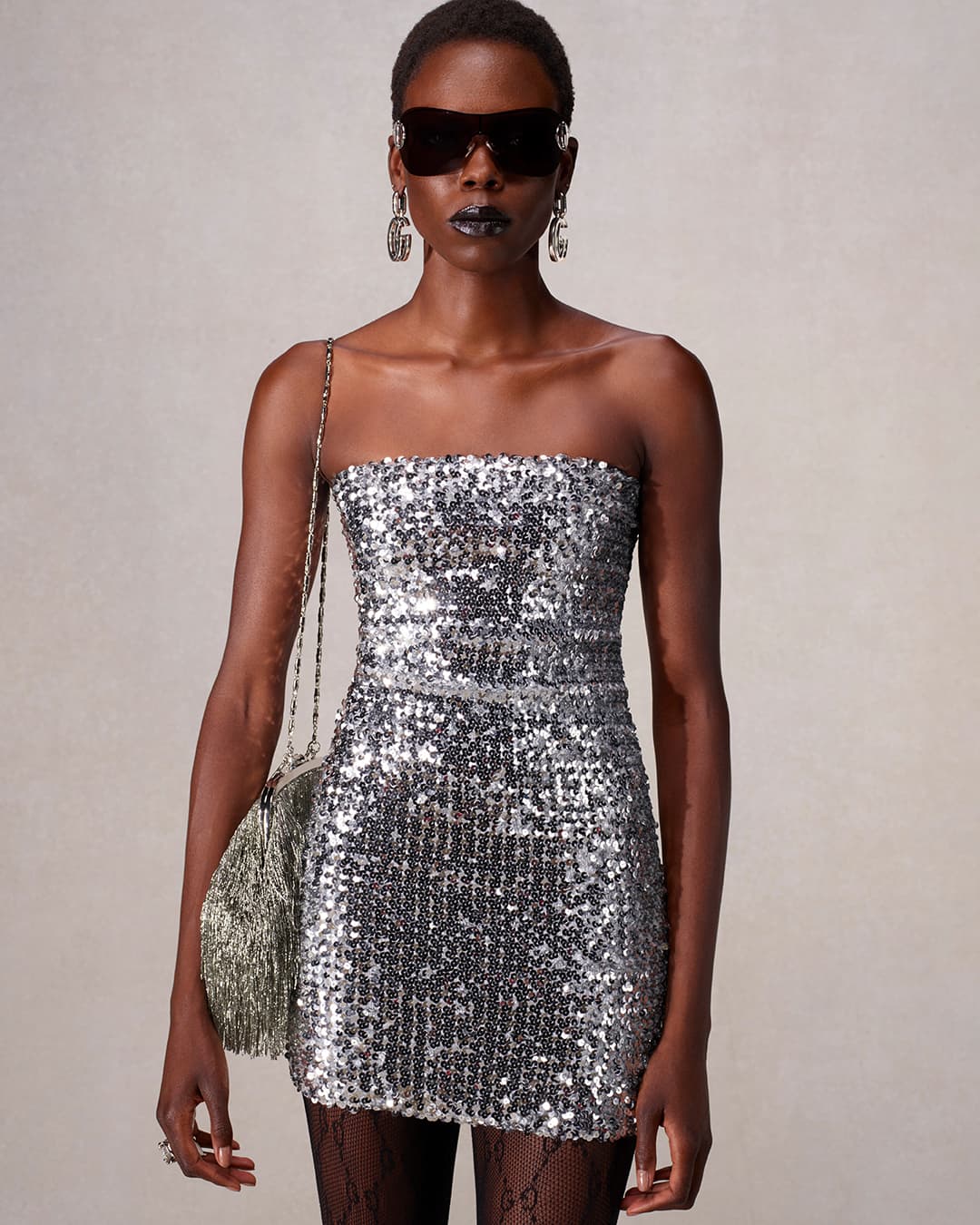Woman in sequin dress with sunglasses and fringe purse. Dark lipstick and black tights with logo. Studio lighting on neutral backdrop.