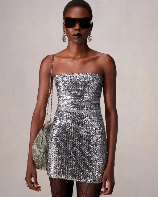 Woman in sequin dress with sunglasses and fringe purse. Dark lipstick and black tights with logo. Studio lighting on neutral backdrop.