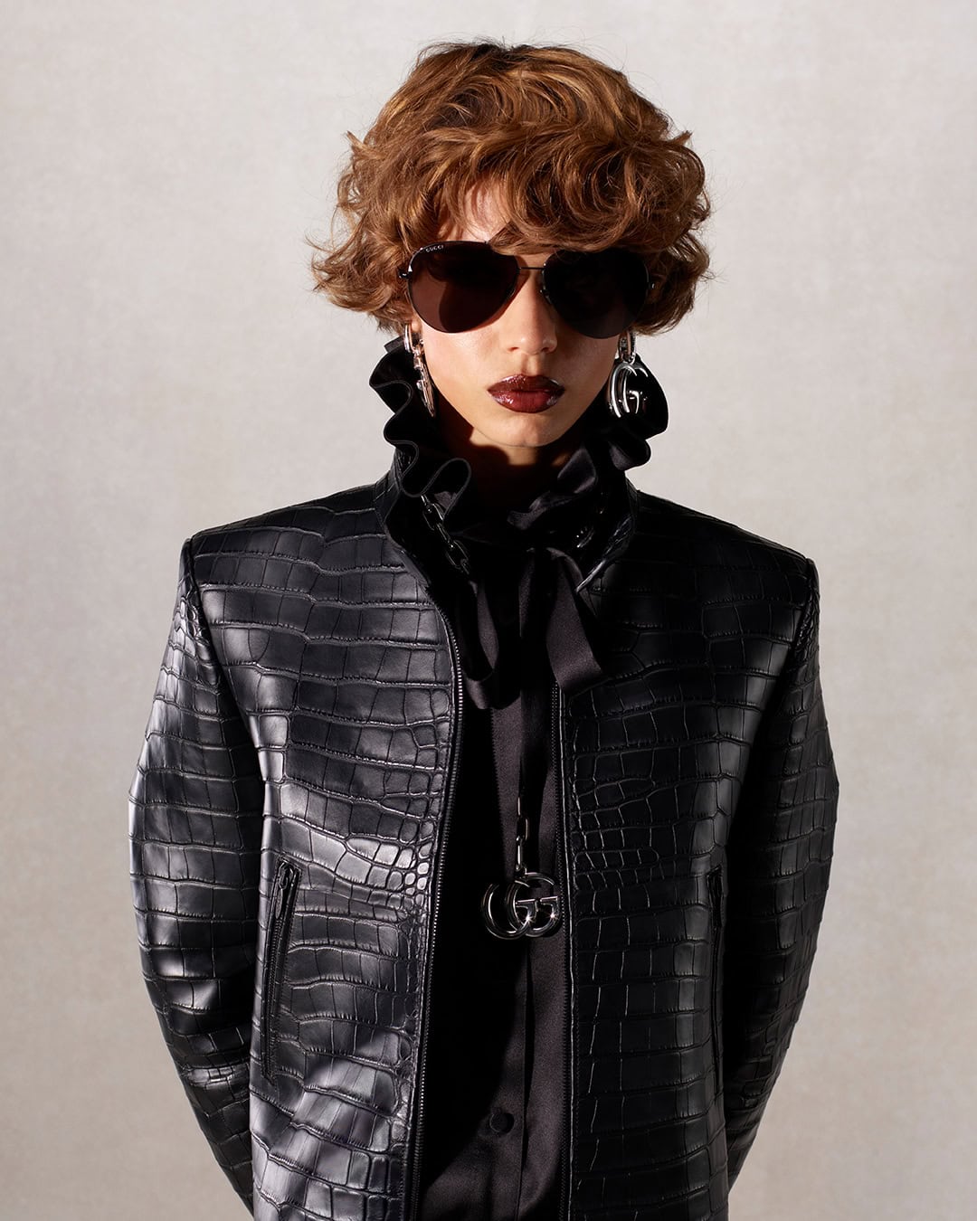 Person wearing sunglasses and a black reptile-patterned leather jacket with a ruffled collar, posing in front of a neutral background.