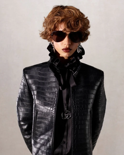 Person wearing sunglasses and a black reptile-patterned leather jacket with a ruffled collar, posing in front of a neutral background.
