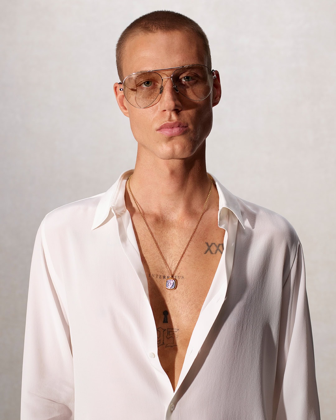 Portrait of a fair-skinned adult male with aviator glasses, gold chain necklace, and unbuttoned white shirt, studio setting.