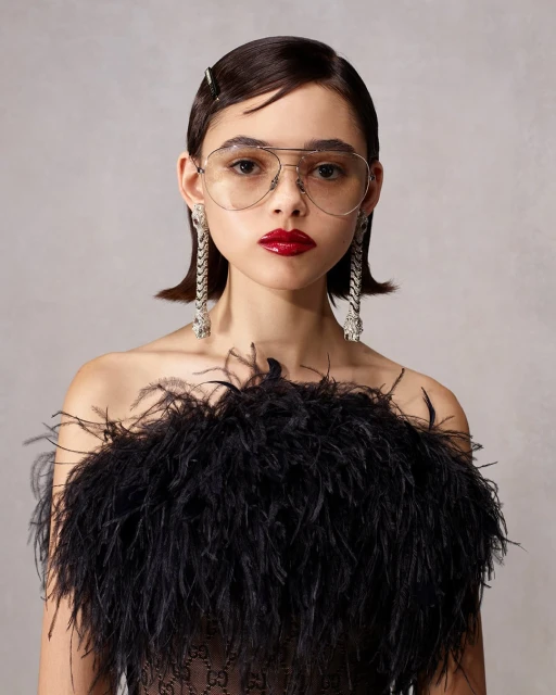 Close-up studio portrait of a young woman with dark hair and aviator glasses wearing a feather-embellished dress, statement earrings, and red lipstick.